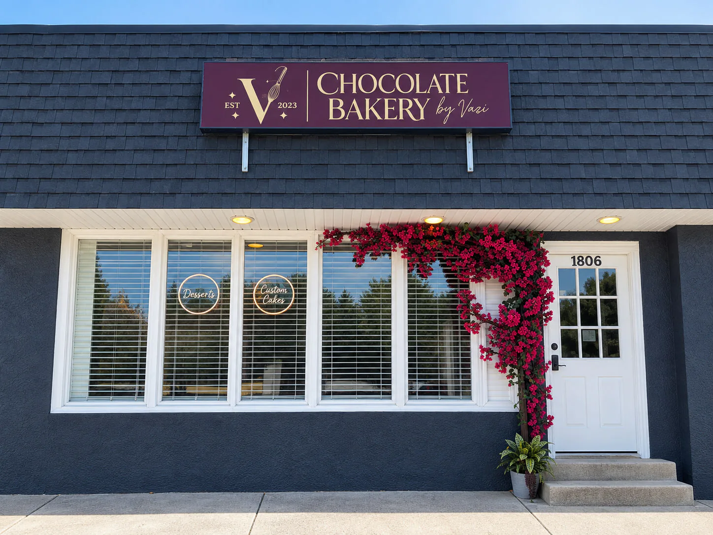 The bakery from the street — a dark shingled storefront under a burgundy sign lettered Chocolate Bakery by Vazi in gold, with a flowering vine climbing beside the white door at 1806