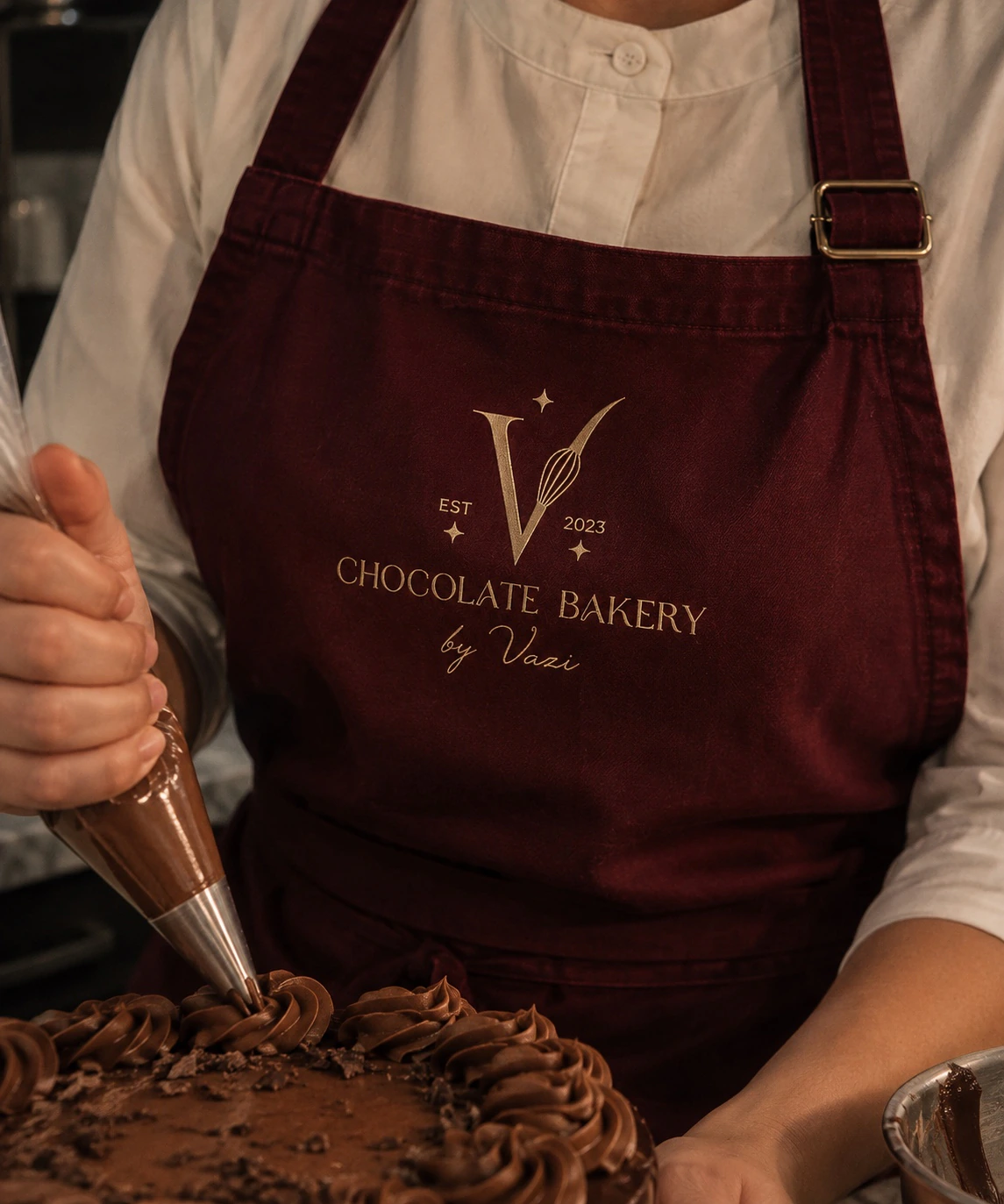 A baker in a burgundy Chocolate Bakery by Vazi apron piping rosettes onto a chocolate cake